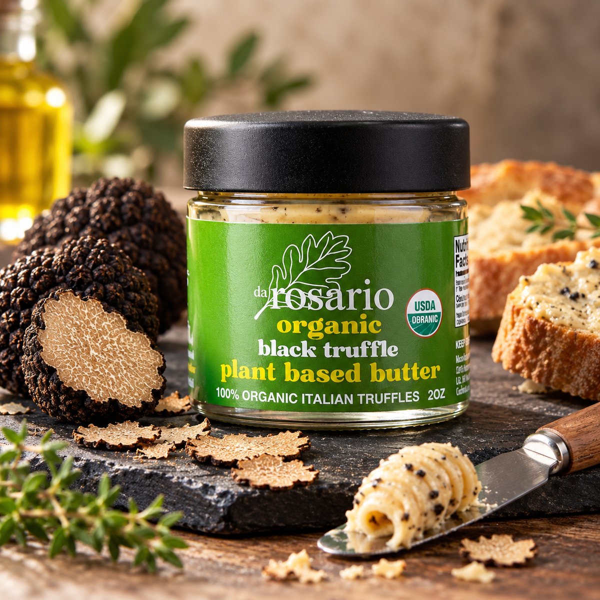 A jar of daRosario organic black truffle plant-based butter on a dark stone board with fresh and sliced black truffles, bread, herbs and buttered toast