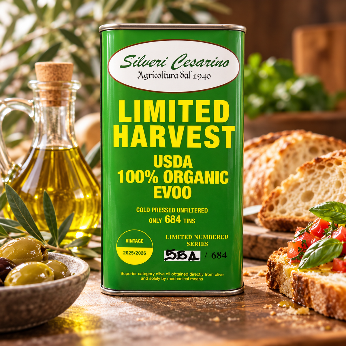 A green can of Silveri Cesarino Organic Evoo olive oil stands on a wooden table surrounded by olives, a glass bottle of oil, bread, and bruschetta. The can features yellow and white text reading 'LIMITED HARVEST USDA 100% ORGANIC EVOO'.