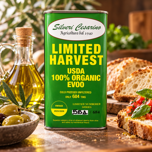 A green can of Silveri Cesarino Organic Evoo olive oil stands on a wooden table surrounded by olives, a glass bottle of oil, bread, and bruschetta. The can features yellow and white text reading 'LIMITED HARVEST USDA 100% ORGANIC EVOO'.