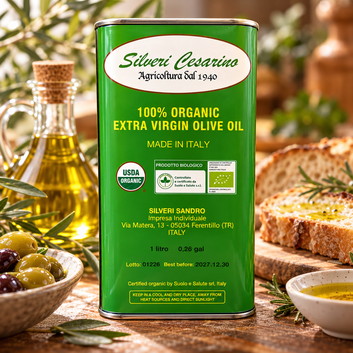 A green canister of Silveri Cesarino 100% Organic Extra Virgin Olive Oil stands on a rustic wooden table, surrounded by olive branches, a glass bottle of oil, olives, and sliced bread.